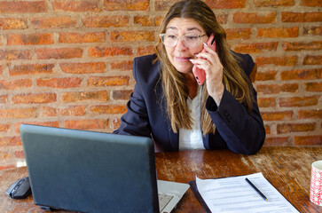 Mature woman working with a laptop and having a conversation on phone.Woman taking notes while talking on mobile phone.