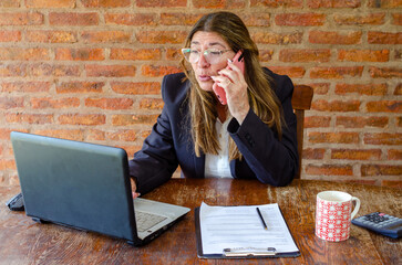 Mature woman working with a laptop and having a conversation on phone.Woman taking notes while talking on mobile phone.