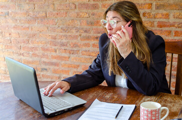 Mature woman working with a laptop and having a conversation on phone.Woman taking notes while talking on mobile phone.