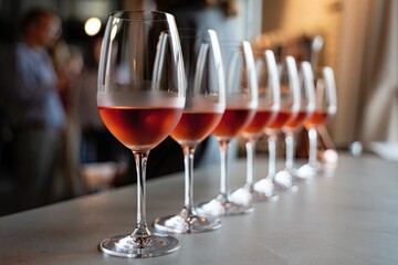 Wine glasses filled with rose or red wine standing in row on bar counter during wine tasting event, with people talkig, evaluating and drinking in background.