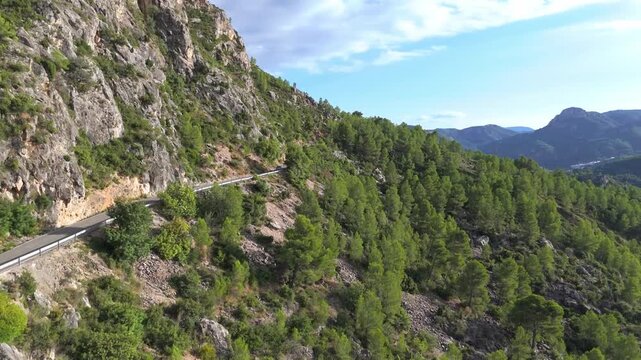 Overhead perspective of a white vehicle navigating a serpentine road etched into a rugged mountainside, adorned with lush pine trees