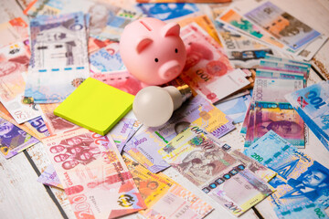 A pink piggy bank, a LED lightbulb, and a yellow sticky note sit on a large, scattered collection of diverse international banknotes, including Angolan Kwanza and Ghanaian Cedis. Global Savings.