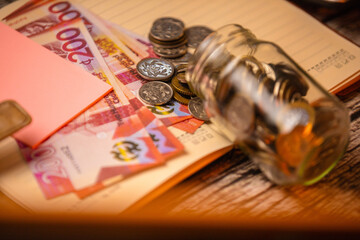 An eye-level shot captures a glass jar of Ghanaian Cedi coins spilling onto a notebook, with 200 cedi notes, a pink sticky note, and a blurred background, highlighting a financial scene
