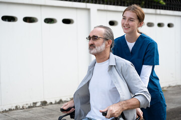 Young caregiver assisting elderly man in wheelchair during outdoor walk. Concept of home healthcare, mobility support, independence, and compassionate senior care in a calm residential area.