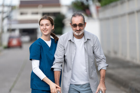 Smiling young caregiver assisting senior man with walker during outdoor walk. Elderly home care, mobility support, rehabilitation, and companion service promoting independence and health.