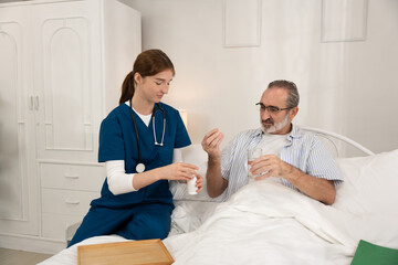 Fototapeta premium Nurse preparing medication for elderly man at home while he holds a glass of water in bed. Concept for home healthcare, medical support, and elderly assistance.