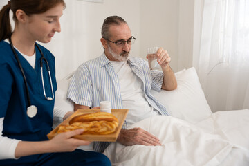 Female nurse serving breakfast and medication to senior man in bed while he drinks water. Ideal for home healthcare, elderly support, daily care routine, and medical service visuals.
