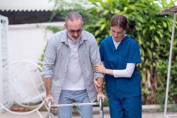 Senior man using a walker assisted by a female nurse outdoors. Concept of elderly care, physical therapy, rehabilitation, recovery support, or mobility assistance for seniors.