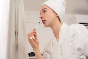 Woman brushing her teeth in bathroom after shower, wearing white bathrobe and towel on head. Morning dental care, oral hygiene, healthy lifestyle routine. brushing, teeth, dental, oral, hygiene, woman