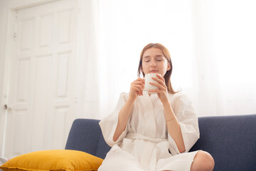 Young woman in bathrobe sitting on sofa with coffee mug, enjoying peaceful morning at home. Calm lifestyle, self-care routine, and cozy indoor relaxation with natural light and positive mindset.