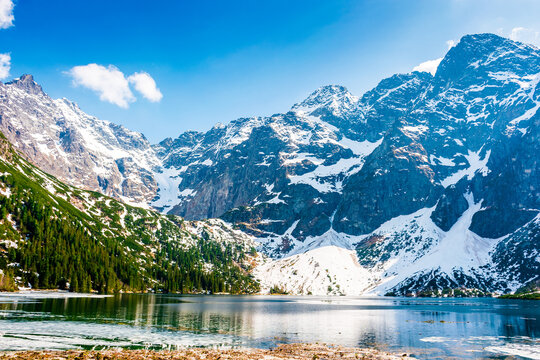 mountain lake in tatra national park. morskie oko surrounded with rugged snow capped peaks in early spring. beautiful view of reflection and ice on water. green environment background under blue sky