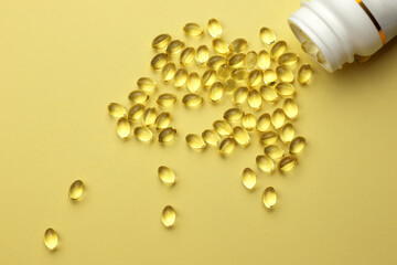 Gelatin capsules with fish oil or vitamin D spilling out of a bottle on a yellow background