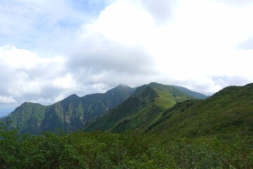 Fototapeta premium Rolling green ridges of Mount Ashibetsu, Hokkaido, bask under shifting clouds, revealing forested slopes and deep valleys across the Yubari Mountains.