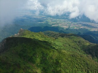 Fototapeta premium Cloud shadows sweep across green ridges on Mount Ashibetsu, Hokkaido, revealing forested slopes and distant patchwork farmland beneath drifting mist.