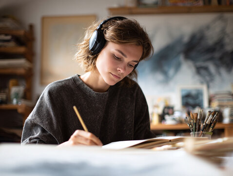 Creative process. Young woman engrossed in drawing, wearing headphones, in a lightfilled, artistic studio. Focus, creativity, art, lifestyle concept.
