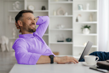 A man sits comfortably at a stylish table, working on his laptop while sipping coffee. Sunlight streams through the window, creating a serene home atmosphere.