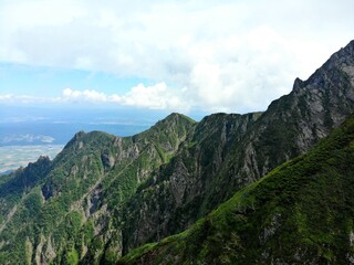 Fototapeta premium Rugged rock ridges carve dramatic cliffs on Mount Ashibetsu, Hokkaido, with green slopes and distant plains under bright cumulus skies.