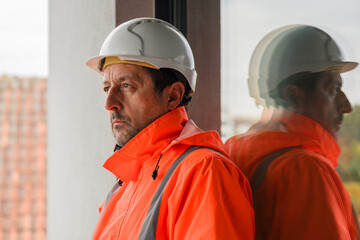 Serious pensive civil engineer in orange safety jacket and white hardhat at construction site, reflection visible on glass window.
