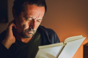 Quiet moment of reflection — mature man absorbed in reading under warm low key light, symbolizing knowledge and introspection.