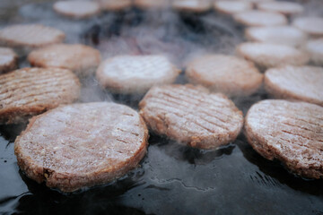 A hand with a ring uses a fork and spatula to flip raw burger patties on a smoky circular grill over glowing charcoal, with flames and steam rising.
