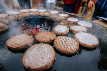 A hand with a ring uses a fork and spatula to flip raw burger patties on a smoky circular grill over glowing charcoal, with flames and steam rising.