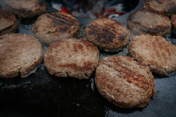 burger patties sizzling on a flat grill above glowing charcoal, with a hand flipping them using fork and spatula. Steam and grill marks visible.
