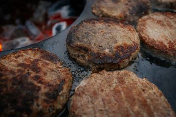 burger patties sizzling on a flat grill above glowing charcoal, with a hand flipping them using fork and spatula. Steam and grill marks visible.