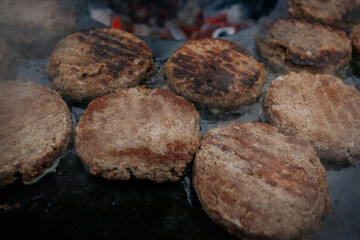 burger patties sizzling on a flat grill above glowing charcoal, with a hand flipping them using fork and spatula. Steam and grill marks visible.
