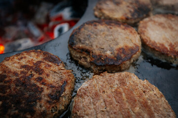 burger patties sizzling on a flat grill above glowing charcoal, with a hand flipping them using fork and spatula. Steam and grill marks visible.
