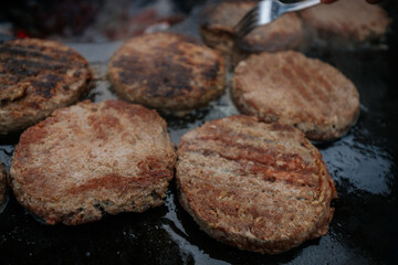 burger patties sizzling on a flat grill above glowing charcoal, with a hand flipping them using fork and spatula. Steam and grill marks visible.