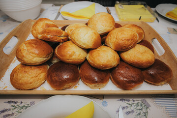 A large pile of golden brown bread rolls arranged on a wooden serving tray, ready to be served at a table set with plates and yellow napkins.