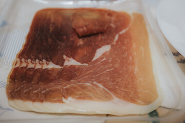 Close-up of thinly sliced prosciutto laid out on a transparent tray, showing marbled texture and rich pink color with white fat edges.