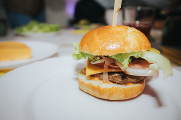 A close-up of a juicy cheeseburger with lettuce, cheddar, onions, and a beef patty in a golden bun, served on a white plate at a lively indoor gathering.