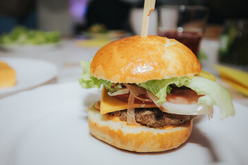 A close-up of a juicy cheeseburger with lettuce, cheddar, onions, and a beef patty in a golden bun, served on a white plate at a lively indoor gathering.