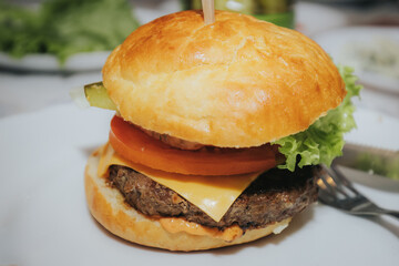 A close-up of a juicy cheeseburger with lettuce, cheddar, onions, and a beef patty in a golden bun, served on a white plate at a lively indoor gathering.