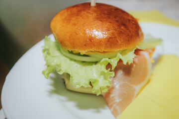 A close-up of a juicy cheeseburger with lettuce, cheddar, onions, and a beef patty in a golden bun, served on a white plate at a lively indoor gathering.