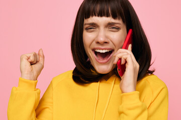 Joyful young woman smiles while talking on a bright red cellphone, wearing a yellow sweater against a pastel pink backdrop, radiating energy, happiness, and casual style.