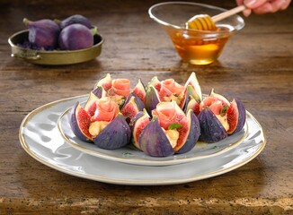Arrangement of halved fresh figs on plates topped with cheese and thin slices of ham. Honey is drizzled using a honey dipper. More fresh figs are in a dish on a wooden table