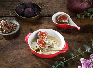 Several dishes showcase fresh figs, pistachios, and a creamy sauce. Gold spoon accents a red and white bowl sitting on a dark wood surface. A delicate floral sprig is placed nearby