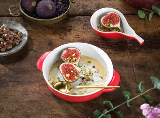 A delightful dessert featuring ripe figs and pistachios sits in a red bowl, golden syrup drizzled on top. The bowl is set on a weathered wooden surface, tempting indulgence