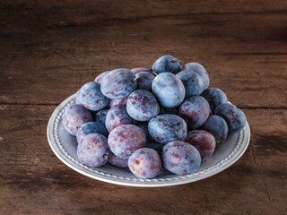 A plentiful collection of ripe plums with a beautiful purple hue are piled high on a light colored plate. The plate sits atop a rustic wooden surface, inviting a taste of summer