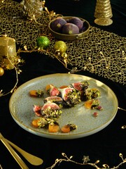 Sliced figs and dried apricots, some coated in chopped pistachios, are presented on a decorative plate, amidst glittering lights, ornaments, and golden cutlery on a dark table