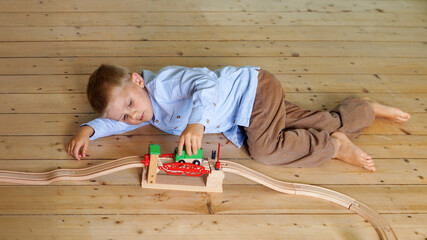 Boy lying on floor moves toy train on wooden railway tracks, balance between loneliness and independence, missing connection. Concept of sustainable childhood, life without screens and modern solitude