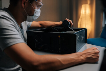 Computer technician wearing protective mask and safety eyewear cleaning desktop tower with handheld vacuum during precision hardware maintenance. Servicing motherboard of dusty computer in workshop. © dikushin