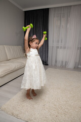 A little girl in a princess dress exercises with dumbbells on TV. Selective focus, real photo.