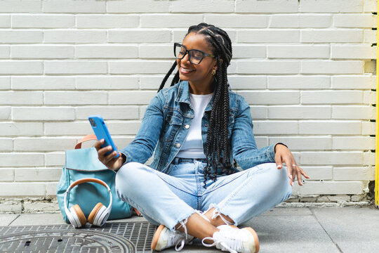 Smiling Latina woman with braided hair Sitting on the street using her smartphone