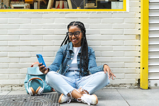 Smiling Latina woman with braided hair Sitting on the street using her smartphone
