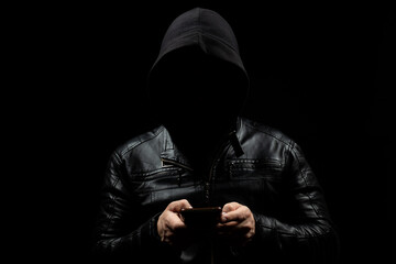 a faceless hooded man holding a smart phone with both hands in the shadows