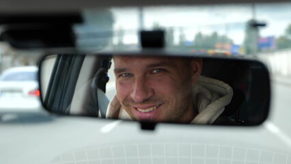 Cheerful adult Caucasian male driving a car in city looking and smiling in rearview mirror. Happy man face reflected in car mirror. Traveling in vehicle. Slow motion concept