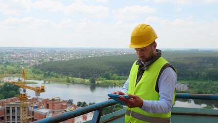 Focused Caucasian young man in uniform and helmet working on digital tablet while standing on construction site. Competent engineer checking working process. Building industry.